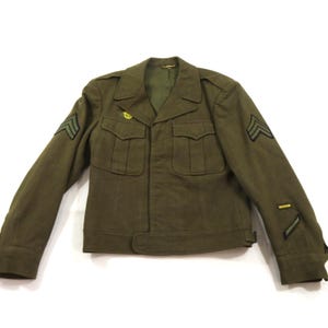 May include: An olive drab military-style jacket featuring a notched collar, two front chest pockets with buttoned flaps, and long sleeves. The sleeves display embroidered chevron rank insignia and service stripes in dark green and black thread.