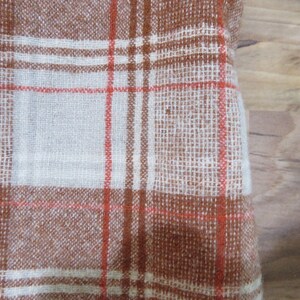 May include: Close-up of a woven fabric with a plaid pattern. The fabric features a brown and white color scheme with thin red lines intersecting to create a grid. The texture appears slightly rough, suggesting a natural fiber.