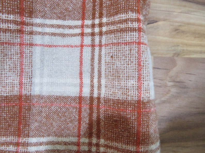 May include: Close-up of a woven fabric with a plaid pattern. The fabric features a brown and white color scheme with thin red lines intersecting to create a grid. The texture appears slightly rough, suggesting a natural fiber.