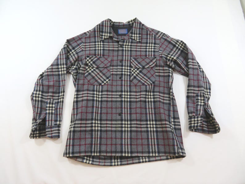 May include: A long-sleeve button-down shirt featuring a grey, black, white, and maroon plaid pattern. The shirt includes a pointed collar, two chest pockets with buttoned flaps, and buttoned cuffs. The Pendleton brand label is visible at the neck.