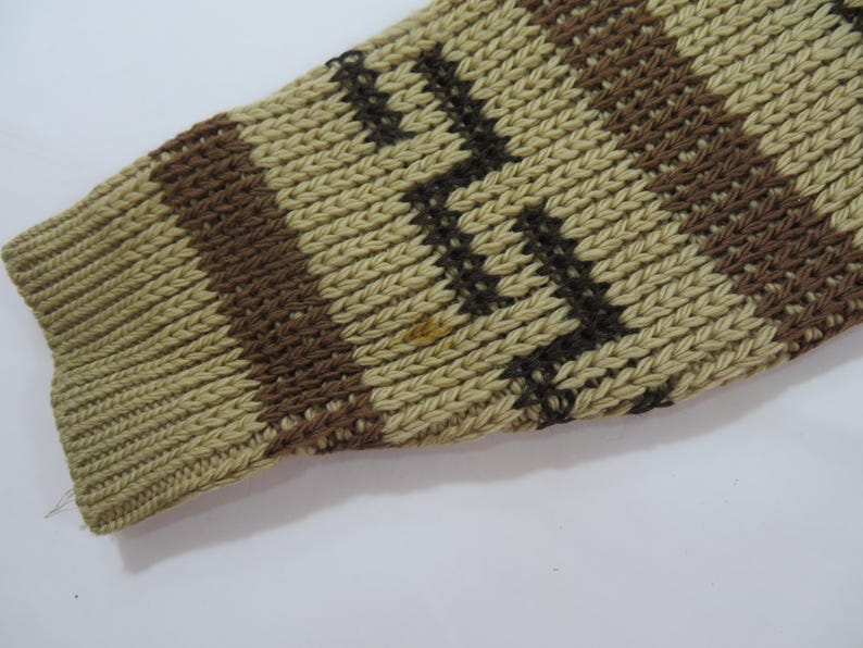 Puede incluir: Primer plano de una manga de punto en tonos beige, marr&oacute;n y negro. La manga presenta un pu&ntilde;o acanalado y un patr&oacute;n geom&eacute;trico de rayas y zigzags. La prenda de punto tiene un aspecto texturizado.