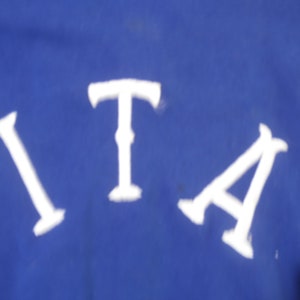 May include: A vibrant royal blue fabric surface featuring the white capital letters I, T, and A arranged in a slight arc. The letters are rendered in a bold, serif-style font with distinct horizontal bars at the top and bottom of each character.