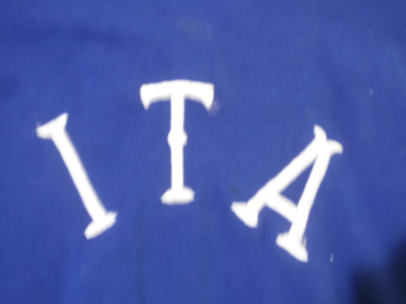 May include: A vibrant royal blue fabric surface featuring the white capital letters I, T, and A arranged in a slight arc. The letters are rendered in a bold, serif-style font with distinct horizontal bars at the top and bottom of each character.