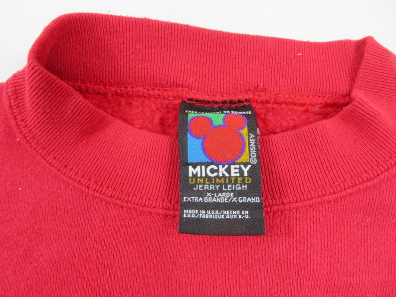 Puede incluir: Primer plano de una sudadera roja con cuello redondo. La etiqueta presenta el logotipo de Mickey Mouse y el texto "MICKEY UNLIMITED JERRY LEIGH X-LARGE EXTRA GRANDE/X GRAND". La etiqueta tambi&eacute;n dice "MADE IN U.S.A."