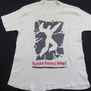 Puede incluir: Camiseta de color crema con un gráfico de una persona rompiendo un fondo gris oscuro y fragmentado. Las palabras "HUMAN RIGHTS NOW!" están impresas en rojo debajo. La camiseta tiene mangas cortas y cuello redondo.