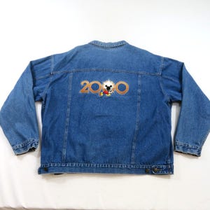 May include: A blue denim jacket featuring a back graphic with the text 2000 in gold lettering. The center of the zero is replaced by a small, colorful illustration of Mickey Mouse with a white starburst effect surrounding the character.