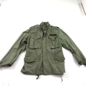 May include: An olive green field jacket featuring a high collar, long sleeves, and four front pockets with buttoned flaps. A black patch with the text U.S. ARMY is positioned above the left chest pocket. The jacket includes a front zip closure.