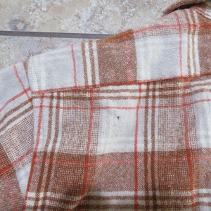 May include: Close-up of a brown, white, and orange plaid flannel shirt. The shirt has a button-down collar and is made of a soft, woven fabric. The plaid pattern is a classic design, suitable for casual wear.