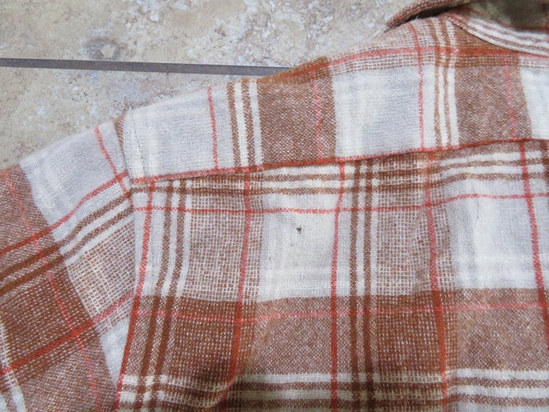 May include: Close-up of a brown, white, and orange plaid flannel shirt. The shirt has a button-down collar and is made of a soft, woven fabric. The plaid pattern is a classic design, suitable for casual wear.