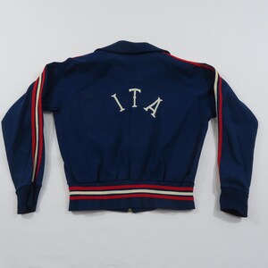 May include: A navy blue zip-up track jacket featuring the letters ITA in white on the back. The sleeves have red and white stripes running down the length, and the hem and cuffs are finished with a ribbed knit trim in red, white, and navy blue.