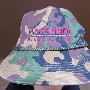 Può includere: Cappello a secchiello mimetico con la scritta "M.R. Tanner Mining, Inc." ricamata in rosa. Il cappello presenta un bordo in corda verde e una tesa. Il tessuto presenta un motivo mimetico verde, marrone e viola.