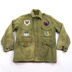 May include: An olive green field jacket featuring a fold-down collar, long sleeves with buttoned cuffs, and four front pockets. The chest area is decorated with four distinct embroidered military-style patches in various shapes and colors.