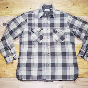 May include: A long-sleeved, button-down shirt with a plaid pattern in shades of gray, white, and red. The shirt has two chest pockets and buttoned cuffs. The shirt is laid flat on a wooden surface.
