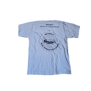 以下が含まれることがあります： 背面に黒いグラフィックがプリントされたライトブルーの半袖Tシャツ。テキストには「PROJECT: SIEGE OF CHARLESTON」、「MOTOROLA」、「MINI-RANGER POSITIONING」と記されており、中央には電子機器のイラストが描かれています。