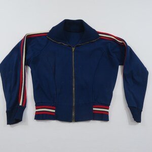 May include: A dark blue zip-up track jacket featuring a high collar and a full-length metal zipper. The sleeves and hem are accented with red and cream-colored stripes. The garment has long sleeves with ribbed cuffs and a ribbed waistband.