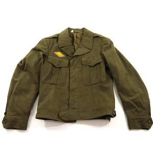 May include: An olive green military-style jacket featuring a collared neckline, long sleeves with buttoned cuffs, and two front chest pockets with flaps. A small, diamond-shaped yellow patch is attached above the right chest pocket.