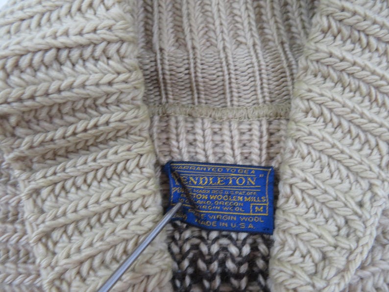 Puede incluir: Primer plano de un su&eacute;ter de punto beige con un patr&oacute;n texturizado. Una etiqueta azul con texto blanco dice "Pendleton" y "Made in U.S.A." El su&eacute;ter es de lana virgen y talla M.