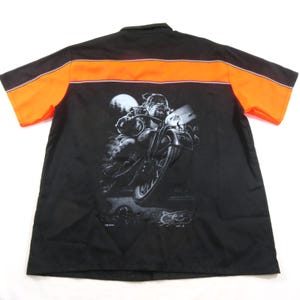 Pode incluir: Uma camisa preta de manga curta com botões e uma faixa horizontal laranja na parte superior das costas e ombros. A parte de trás apresenta um gráfico grande e detalhado em preto e branco de um bulldog montado numa motocicleta sob a lua cheia.