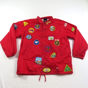 May include: A bright red long-sleeve coach jacket featuring a snap-button front closure, a collar, and numerous colorful embroidered patches across the chest and sleeves. The jacket includes a drawstring hem and the number 431 on the upper sleeve.