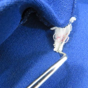 May include: A vibrant royal blue fabric garment featuring a small, frayed white interior tag with faint red markings. A thin, curved silver metal tool is positioned underneath the tag, lifting it slightly away from the textured blue material.