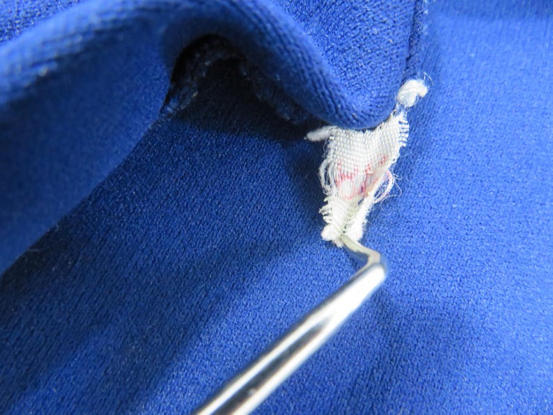 May include: A vibrant royal blue fabric garment featuring a small, frayed white interior tag with faint red markings. A thin, curved silver metal tool is positioned underneath the tag, lifting it slightly away from the textured blue material.