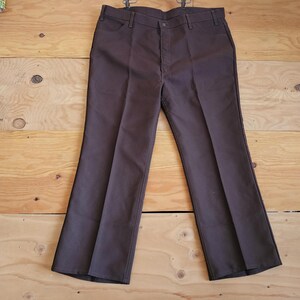 Puede incluir: Un par de pantalones marrones de pierna ancha con un ajuste recto desde la cintura hasta la rodilla y una abertura de pierna acampanada.