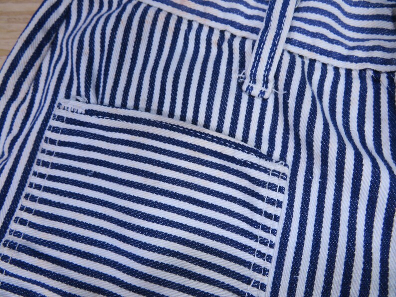 May include: Close-up of blue and white striped fabric with a pocket. The fabric has a woven texture and is sewn with white thread.