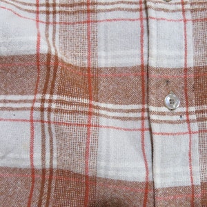 May include: Close-up of a button-down shirt with a plaid pattern. The shirt features a white base with brown and red intersecting lines, creating a classic plaid design. A single button is visible.
