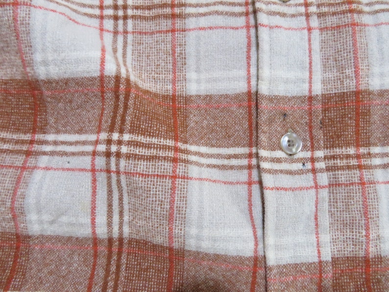 May include: Close-up of a button-down shirt with a plaid pattern. The shirt features a white base with brown and red intersecting lines, creating a classic plaid design. A single button is visible.