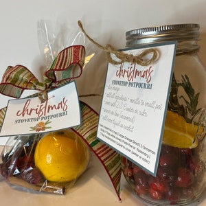 Puede incluir: Dos frascos de vidrio llenos de arándanos, rodajas de naranja, palitos de canela y ramitas de romero. Los frascos están atados con cuerda y una cinta roja y blanca a rayas. Una etiqueta escrita a mano en el frasco dice "Christmas Stovetop Potpourri" e incluye instrucciones de uso.
