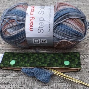 May include: A ball of blue, pink and beige yarn labelled "mary max! Step Soft Superwash Wool/Natur 50g" with a green and beige fabric knitting needle case with two green buttons and two gold knitting needles.