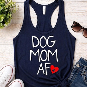 May include: Navy blue tank top with the words "DOG MOM AF" in white, a red heart with a paw print, and a racerback design. The image also includes white sneakers, denim shorts, and sunglasses.