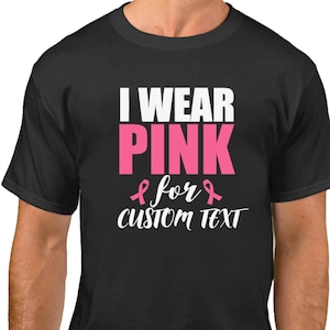 May include: Black t-shirt with the text "I WEAR PINK" in white and pink, with a pink ribbon graphic. Below the text is "for CUSTOM TEXT" in white. The bottom of the image has the text "MANY COLORS" in white on a green background.