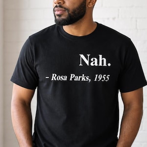 May include: Black t-shirt with the text "Nah. - Rosa Parks, 1955" in white. The shirt is a crew neck style and is being worn by a person. The person is standing near a window and a large green plant.