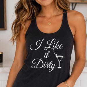 May include: A dark gray tank top with the text "I Like it Dirty" in white script. The tank top has a racerback design and is paired with denim shorts. A cocktail menu is visible in the background.