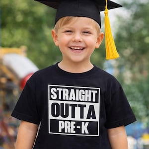 May include: A young child wearing a black graduation cap with a yellow tassel and a black t-shirt. The shirt has the text "STRAIGHT OUTTA PRE-K" in a white box. The child is smiling and wearing khaki pants.