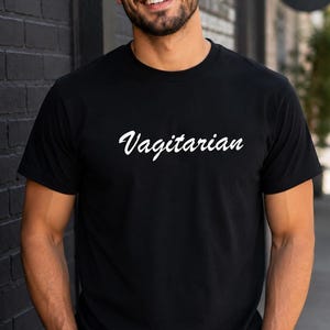 May include: Black t-shirt with the word "Vagitarian" in white script. The shirt is made of a soft material. The person in the image is wearing sunglasses and has a beard. The background includes a black brick wall and a sign that says "Coffee & Good Vibes."