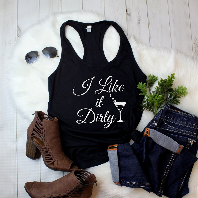 May include: Black racerback tank top with the text "I Like it Dirty" in white script, accompanied by a cocktail glass graphic. The tank top is paired with blue jeans, brown ankle boots, and sunglasses.