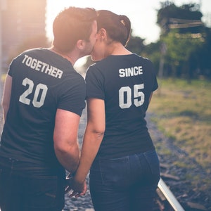 Op de afbeelding: Een stel draagt ​​bijpassende zwarte T-shirts met witte tekst. Het shirt van de man zegt "Together 20" en het shirt van de vrouw zegt "Since 05". Ze houden elkaars hand vast en lopen op een spoorweg.