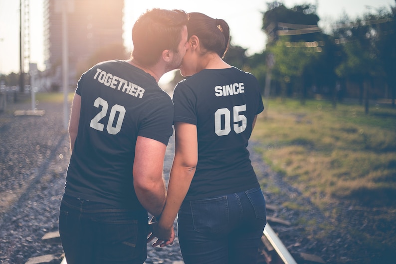 Op de afbeelding: Een stel draagt ​​bijpassende zwarte T-shirts met witte tekst. Het shirt van de man zegt "Together 20" en het shirt van de vrouw zegt "Since 05". Ze houden elkaars hand vast en lopen op een spoorweg.
