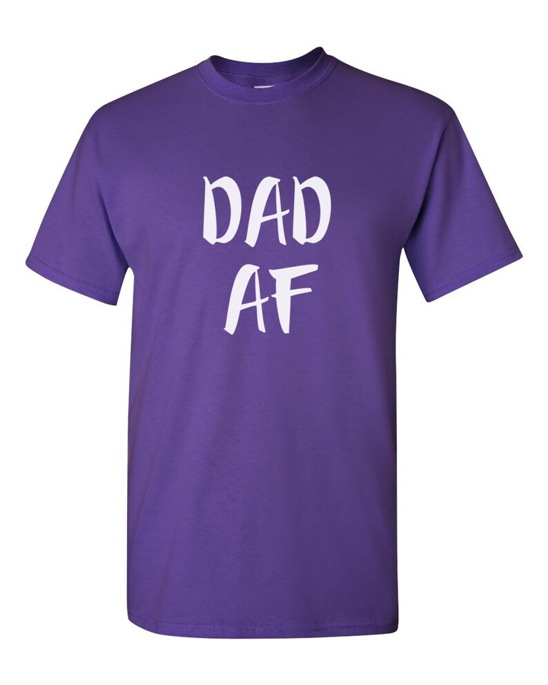 Peut inclure: Un t-shirt violet avec le texte blanc "DAD AF" &eacute;crit &agrave; la main. Le t-shirt a des manches courtes et un col rond.