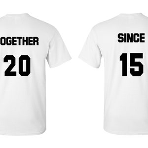 Op de afbeelding: Twee witte T-shirts met zwarte tekst. Het eerste shirt zegt "TOGETHER 20" en het tweede shirt zegt "SINCE 15".