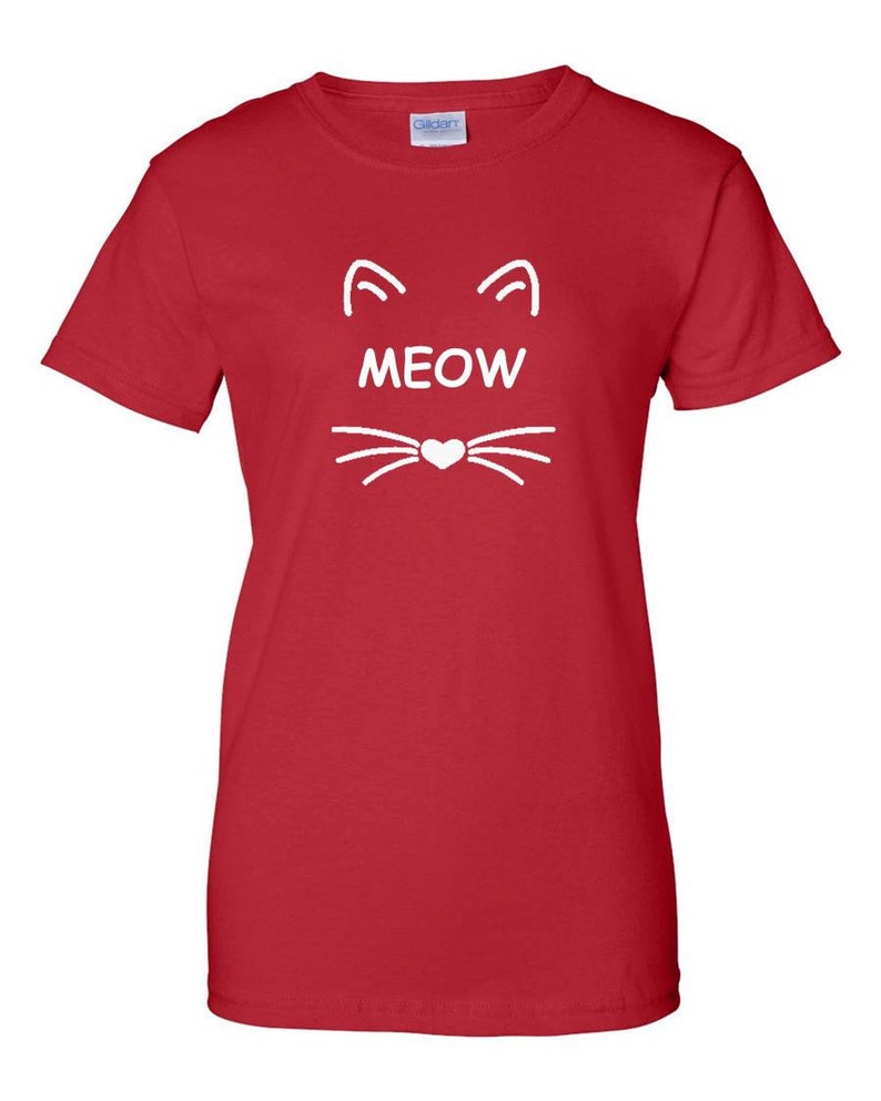 Puede incluir: Camiseta roja con un dise&ntilde;o de cara de gato blanco. El dise&ntilde;o incluye orejas de gato, la palabra "MEOW" y bigotes con una nariz en forma de coraz&oacute;n. La camiseta est&aacute; hecha de un material suave.