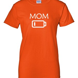 Pu&ograve; includere: T-shirt arancione con la scritta "MOM" in lettere maiuscole bianche sopra un'icona di batteria bianca con un indicatore di batteria scarica. La maglietta &egrave; in materiale morbido e ha maniche corte.