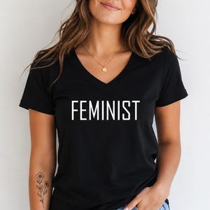 May include: A black short-sleeved t-shirt with a V-neckline featuring the word FEMINIST printed in white capital letters across the chest. The garment is paired with blue denim jeans, creating a casual look with a clear, bold graphic design.