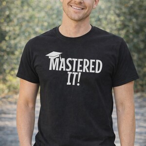K&ouml;nnte beinhalten: Schwarzes T-Shirt mit dem wei&szlig;en Text "MASTERED IT!" und einer Abschlusskappe. Ein lila Banner in der oberen rechten Ecke zeigt "MASTERS DEGREE". Die Person l&auml;chelt und tr&auml;gt das T-Shirt.