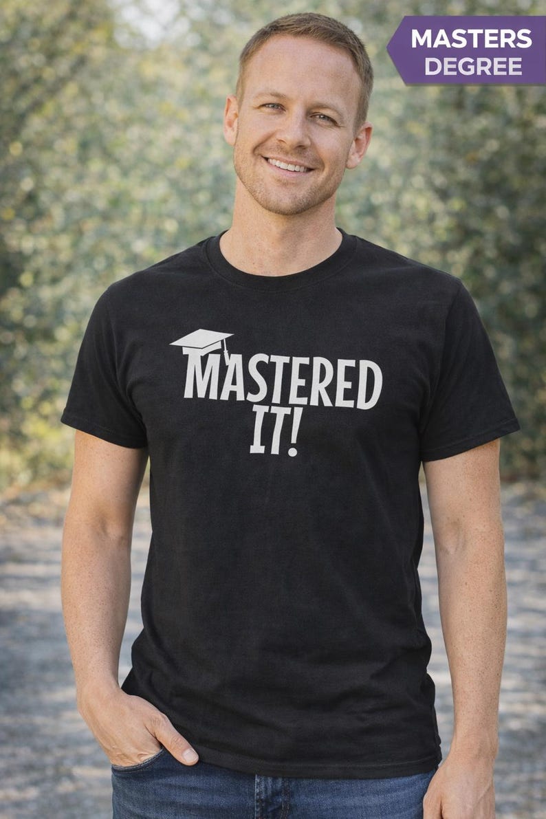 K&ouml;nnte beinhalten: Schwarzes T-Shirt mit dem wei&szlig;en Text "MASTERED IT!" und einer Abschlusskappe. Ein lila Banner in der oberen rechten Ecke zeigt "MASTERS DEGREE". Die Person l&auml;chelt und tr&auml;gt das T-Shirt.