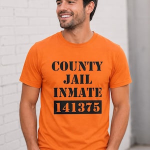 May include: Orange t-shirt with the words "COUNTY JAIL INMATE" printed in black, along with the number "141375". The shirt is made of a soft material and has short sleeves.