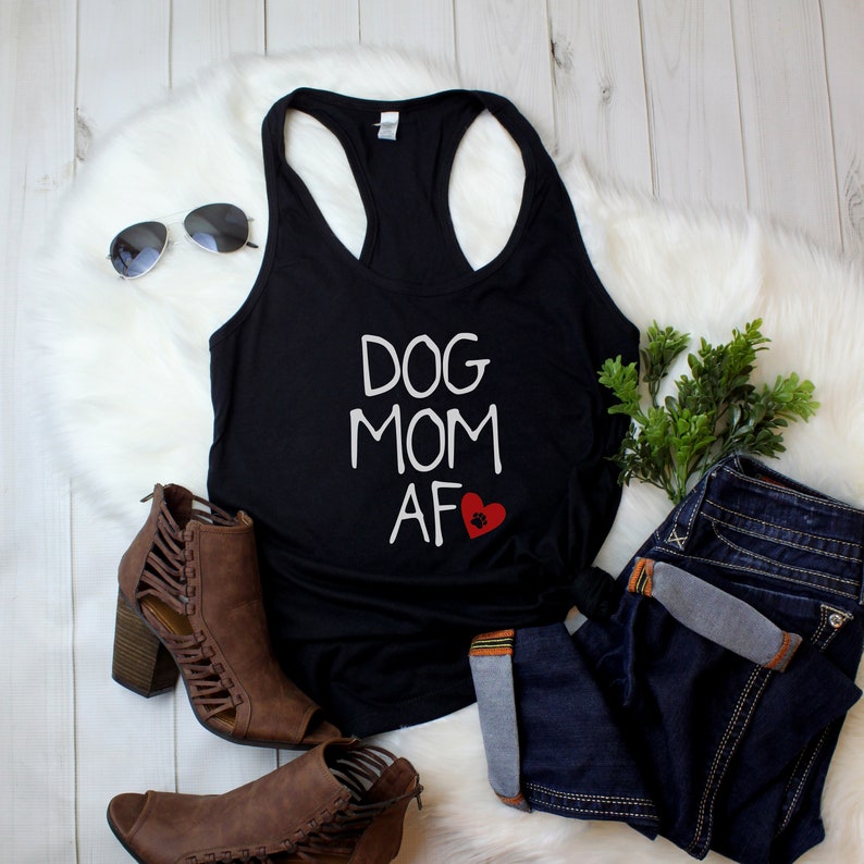 May include: Black racerback tank top with the text "DOG MOM AF" in white, a red heart, and a paw print. The tank top is paired with blue jeans, brown ankle boots, and sunglasses.
