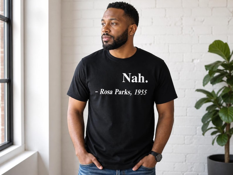 May include: Black t-shirt with the text "Nah. - Rosa Parks, 1955" in white. The shirt is a crew neck style and is being worn by a person. The person is standing near a window and a large green plant.